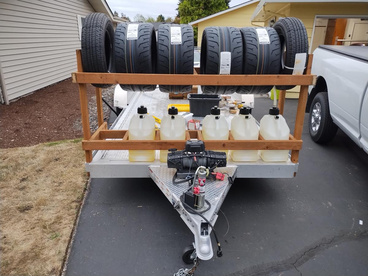 An 18' aluminum open car hauling trailer with a home made rack made of pressure treated plywood with tires and fuel on the front of it.